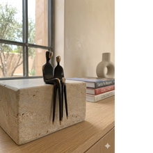 Load image into Gallery viewer, A highly sophisticated, close-up fine art photograph of the authentic 4.5-inch 'The Two of Us bronze sculpture by Yenny Cocq, featuring the two seated abstract figures with dark patina bodies and polished golden-bronze heads and pin-like feet. The figures are perched on the edge of block of polished cross-cut travertine. The unique, extremely elongated legs hang gracefully and securely suspended over the edge of the stone or shelf edge.