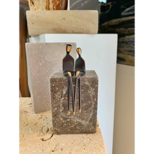 Load image into Gallery viewer, A clean, high-resolution product photograph of 'The Two of Us' (300-12) 4.5-inch miniature bronze sculpture by Santa Fe artist Yenny Cocq. The two abstract, seated figures are perched on a block of polished cross-cut travertine stone against a neutral gray studio background. Their signature elongated legs hang gracefully over the edge. The figures show the characteristic dark, hand-rubbed patina with bright, polished golden-bronze heads that catch the focused gallery lighting. This shot isolates the unique